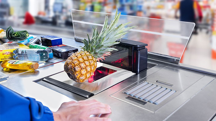 Ananas leżący na szybie skanera dwupłaszczyznowego Datalogic wbudowanego w ladę kasową, obok taśma z zakupami