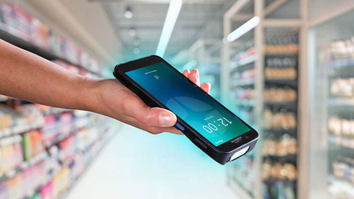 Dłoń trzymająca smukły terminal Datalogic Memor na alejce sklepowej, widoczny profil obudowy i okno skanera na dolnej krawędzi