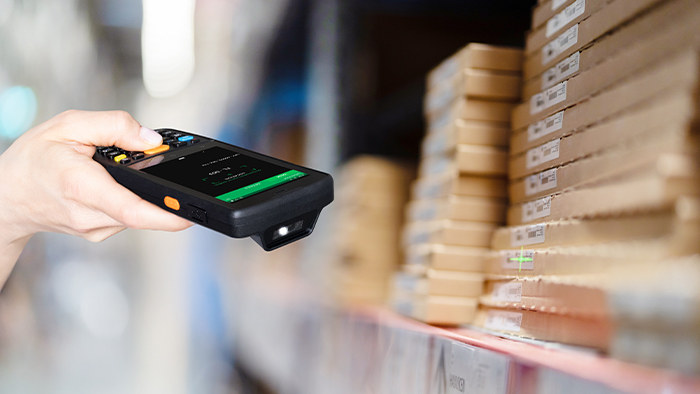 Dłoń trzymająca terminal Datalogic Memor K z klawiaturą numeryczną przy regale z ułożonymi kartonami w magazynie