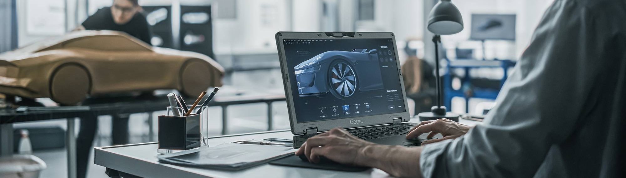 Projektant pracuje na wzmocnionym laptopie Getac S510AD w studiu projektowym samochodów, obok gliniany model auta