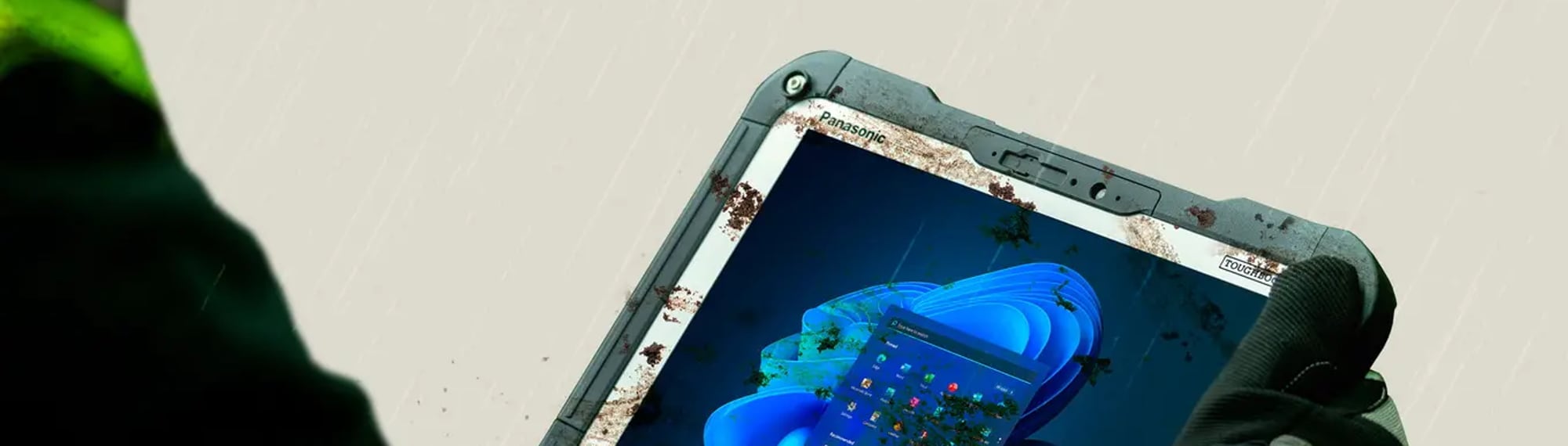 Pracownik w rękawicach trzyma zabłocony wzmocniony tablet Panasonic TOUGHBOOK G2 podczas pracy w deszczu