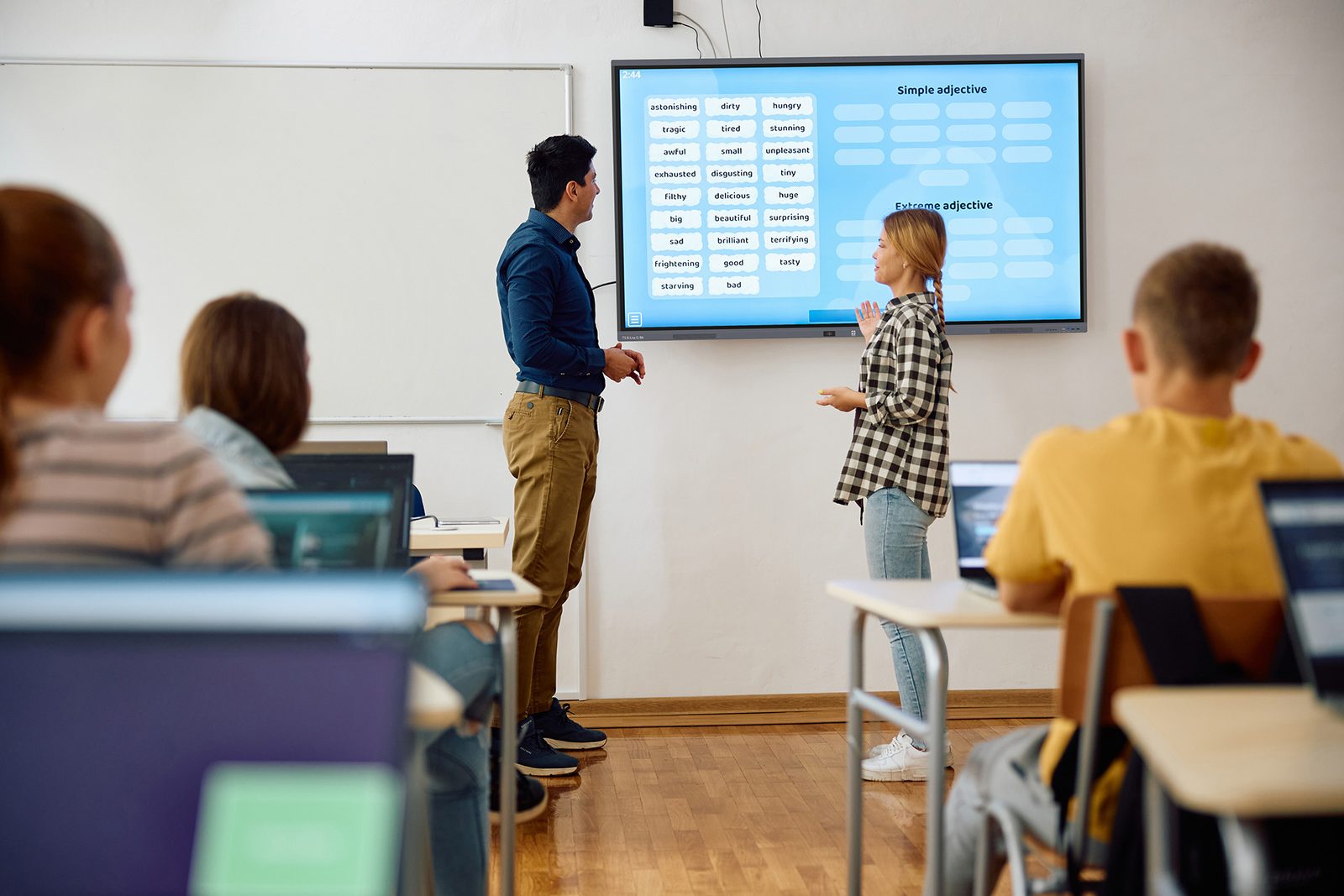 Nauczyciel prowadzi lekcję przy monitorze interaktywnym Avtek TS 8 Lite G, uczennica wskazuje element na ekranie, uczniowie przy ławkach z laptopami